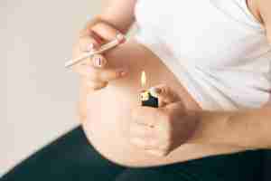 Selective focus of lighter and cigarette in hands of pregnant woman. Female in white shirt smoking during pregnancy period.
